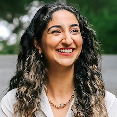 A woman with long curly hair outdoors with a big smile on her face.