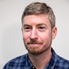A white man with short hair and a bit of a ginger beard with a twinkle in his eye, wearing a plaid shirt.