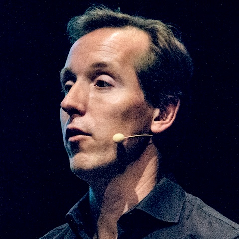 A dramatically lit close-up of a man speaking on stage.