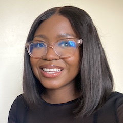 The smiling face of a young black woman with straight shoulder-length dark hair and glasses against a light background.